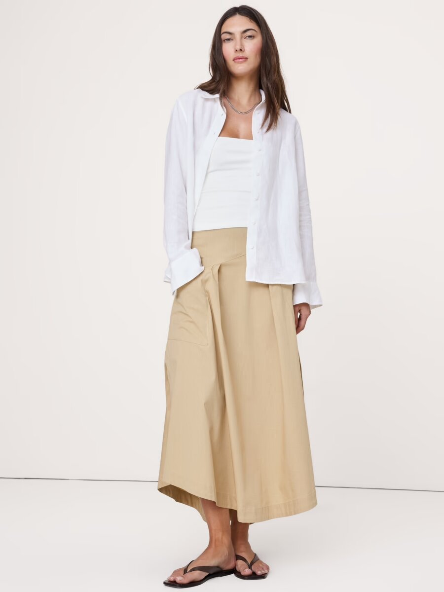 Woman wearing a white button-up shirt, white top, beige midi skirt, and black sandals, standing against a plain light background—a timeless look that showcases effortless fashion and classic style tips.
