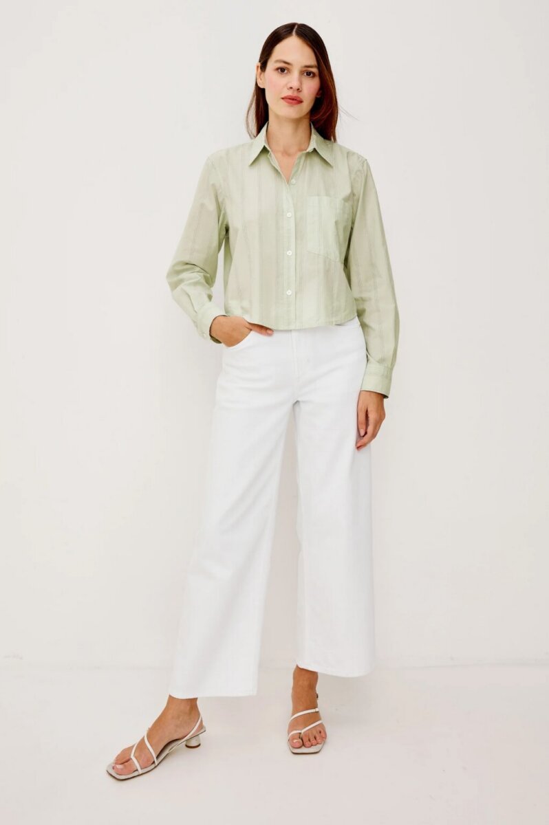 Person standing against a plain background, wearing a light green style button-down shirt, white wide-leg pants, and white sandals, with one hand in a pocket.