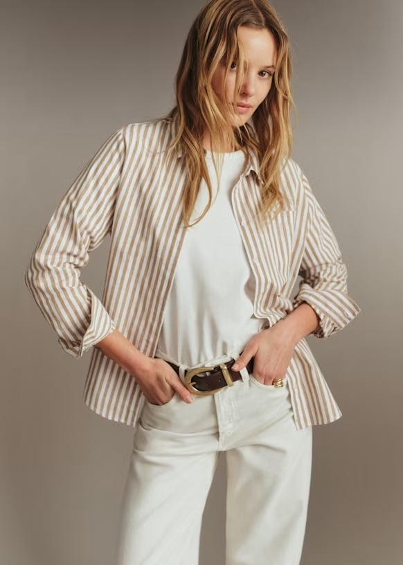 A person stands with hands in pockets, showcasing effortless style in a beige and white striped button-down shirt over a white t-shirt, cream pants, and a brown belt, against a neutral background.