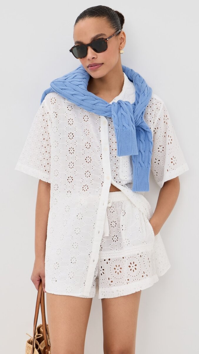 Woman wearing a white eyelet button-down shirt and shorts set, with a blue sweater draped over her shoulders, sunglasses, and carrying a woven bag—an effortlessly chic look that exudes timeless style.