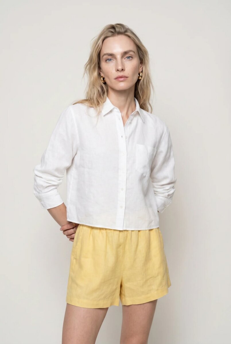 A person stands against a plain background, wearing a white button-up shirt with long sleeves and yellow shorts, looking directly at the camera—an effortlessly fresh example for anyone seeking button-down outfit ideas.