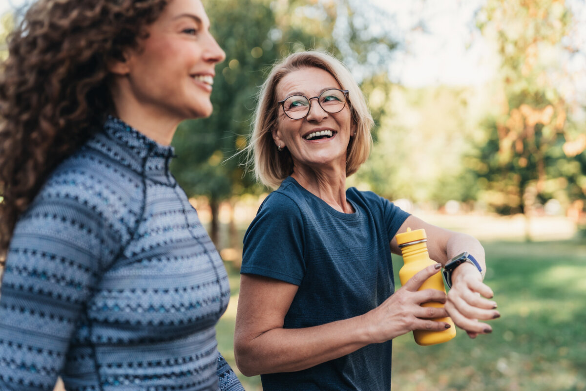 Two women are walking outdoors in a park, embracing an active lifestyle that supports women's healthcare. One holds a yellow water bottle and wears glasses, while both are smiling and dressed in activewear. Trees and greenery are visible in the background.