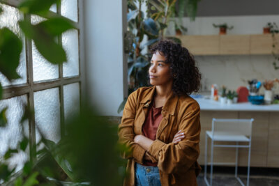 A woman with curly hair stands in a kitchen with arms crossed, looking out a frosted window surrounded by green plants.