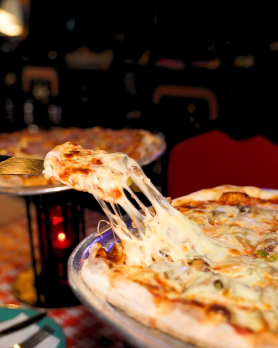 A slice of pizza being lifted from a pan, with melted cheese stretching, in a restaurant setting.