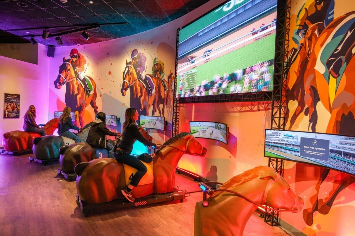People ride mechanical horse simulators facing screens that display virtual horse races in a brightly lit, horse-themed arcade setting—a hidden gem you’ll want to add to any I-65 road trip travel guide.