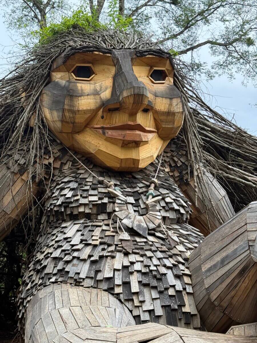 Large wooden sculpture of a troll with a textured, shingled body and twig hair, set outdoors among trees—an unexpected roadside gem if you’re planning a road trip along I-65.