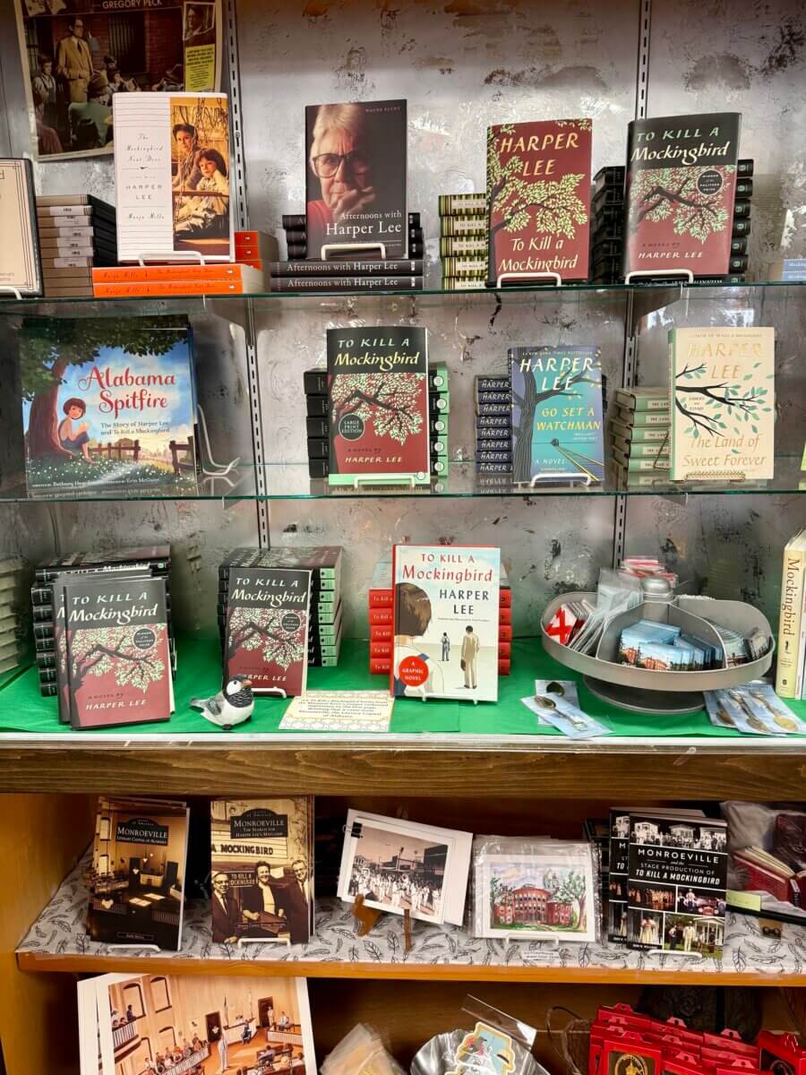 Books and souvenirs related to Harper Lee and "To Kill a Mockingbird" are displayed on glass shelves—including various editions, postcards, and memorabilia.