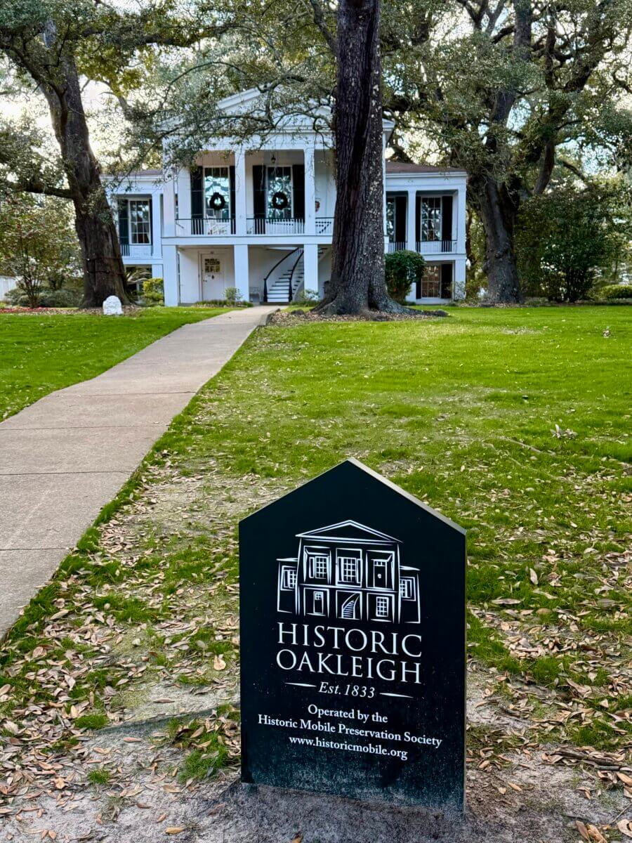 A sign for Historic Oakleigh stands on a lawn, with a white, two-story historic house and large trees in the background—an inviting stop.