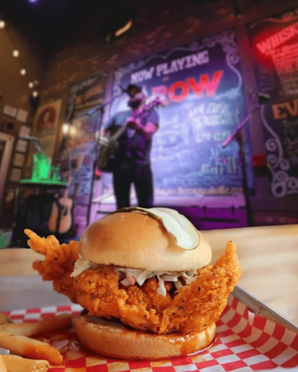 Fried chicken sandwich with sauce and coleslaw in the foreground, perfect for refueling during an I-65 road trip, with a musician performing on stage in a dimly lit restaurant or bar.