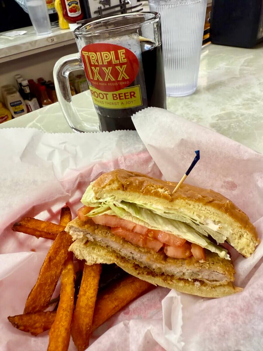 A breaded chicken sandwich with lettuce and tomato, sweet potato fries, a glass of root beer, and a water on a diner counter—the perfect fuel for your Ultimate Road Trip along I-65.