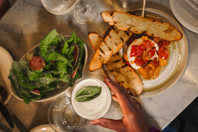 A hand holds a cocktail with a basil leaf beside a plate of grilled bread, burrata with tomatoes, and a bowl of green salad on a marble table—capturing the vibrant flavors and stylish ambiance found in the best Oxford restaurants.