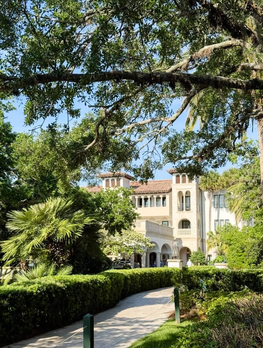 8 Luxurious, Family-Friendly Resorts in the South - 10 A Mediterranean-style building with arched doorways and palm trees is partially shaded by large oak branches along a stone pathway, creating an inviting atmosphere perfect for Southern families seeking memorable family vacations.