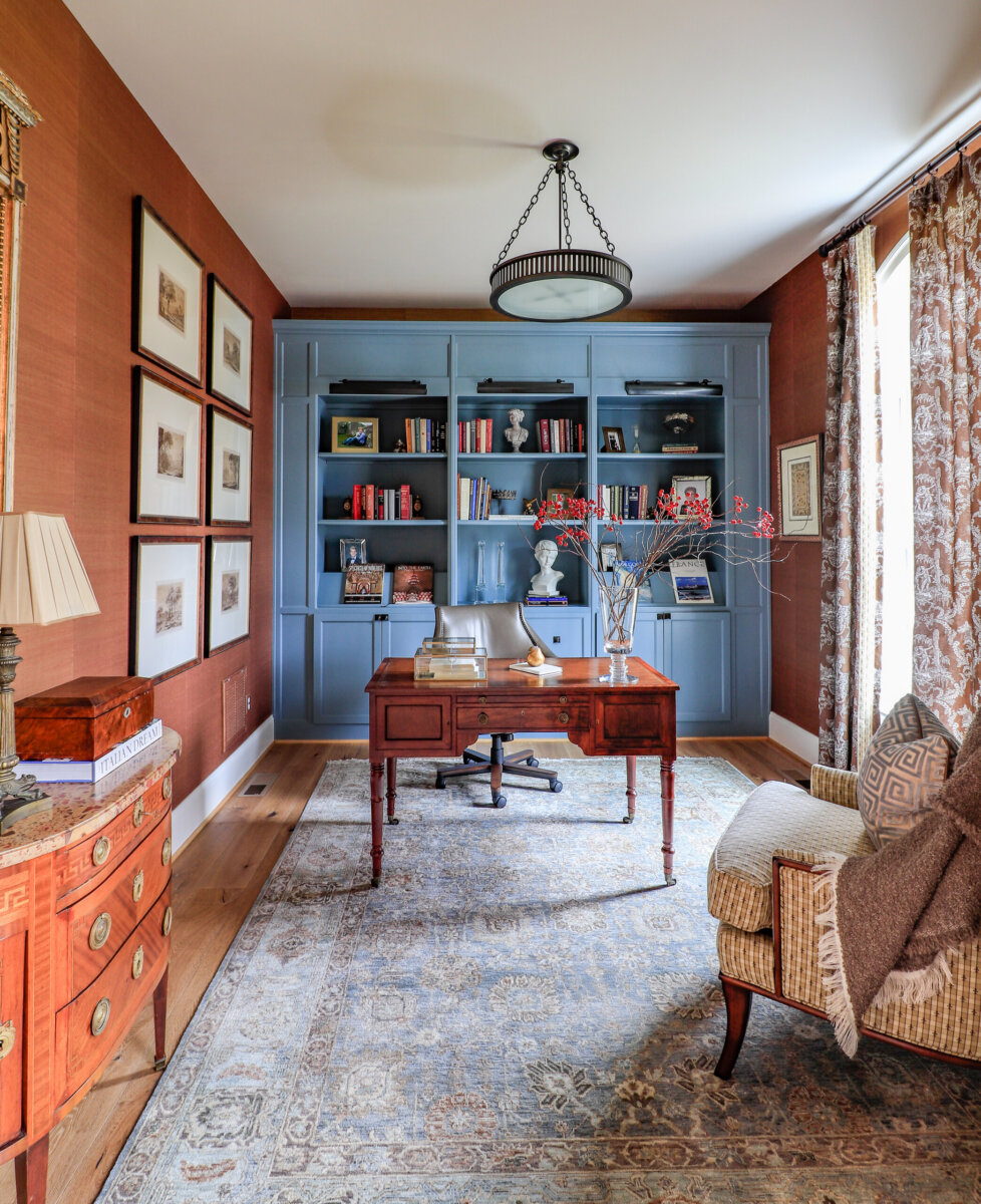 This Designer Wrote the Book on Creating Colorful Spaces — Literally! - 9 A traditional home office with a designer wooden desk, blue built-in bookshelves, an upholstered chair, framed art on the wall, and patterned curtains by a large window.