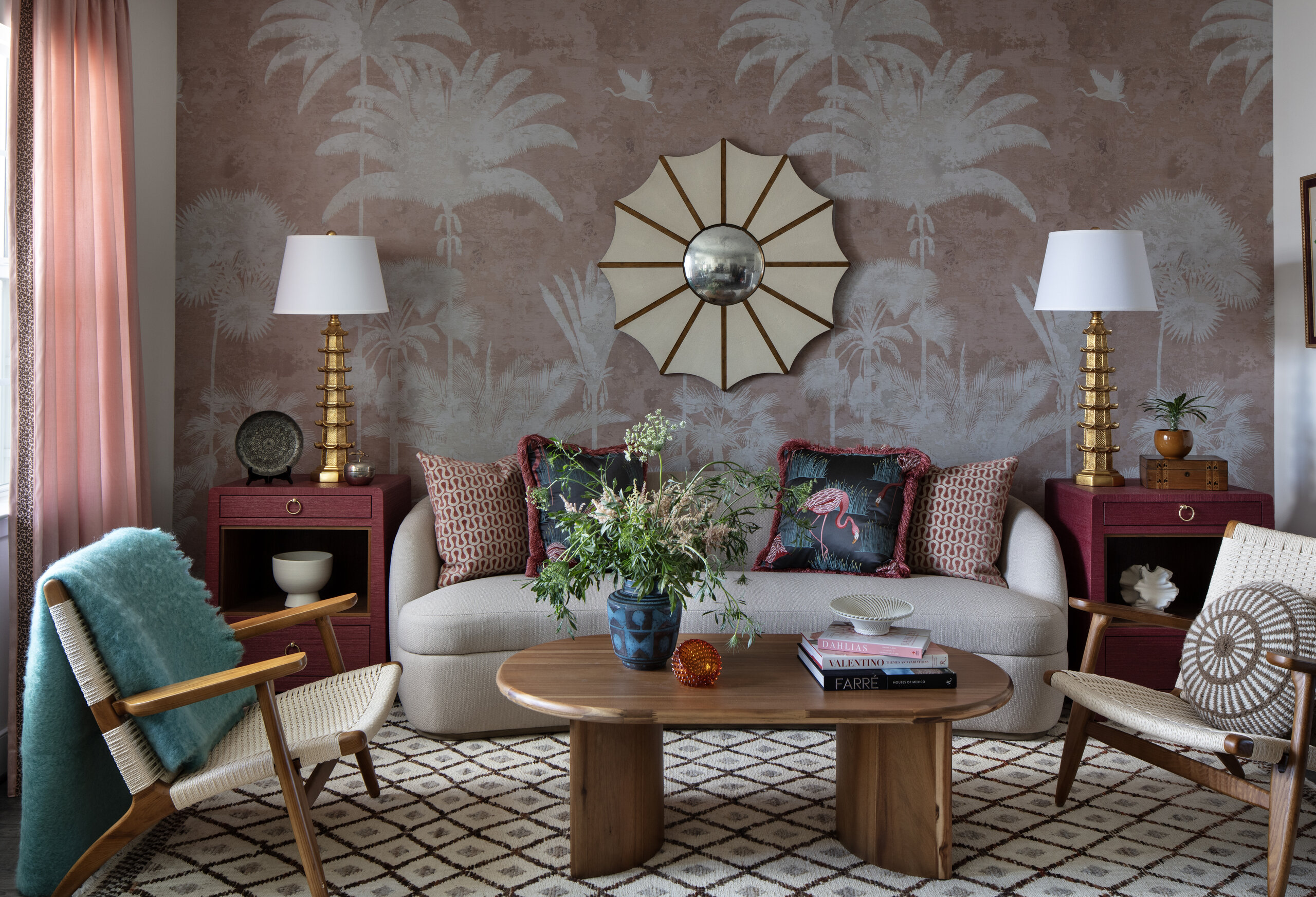A living room with a beige sofa, patterned pillows, two gold lamps, a round sunburst mirror, wooden coffee table, armchairs, and palm-patterned wallpaper.