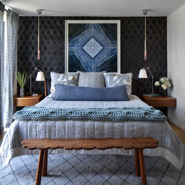 This Designer Wrote the Book on Creating Colorful Spaces — Literally! - 3 A modern bedroom with a patterned black accent wall, large artwork above the bed, blue and gray bedding, wooden nightstands with a designer book, and a wooden bench at the foot of the bed.