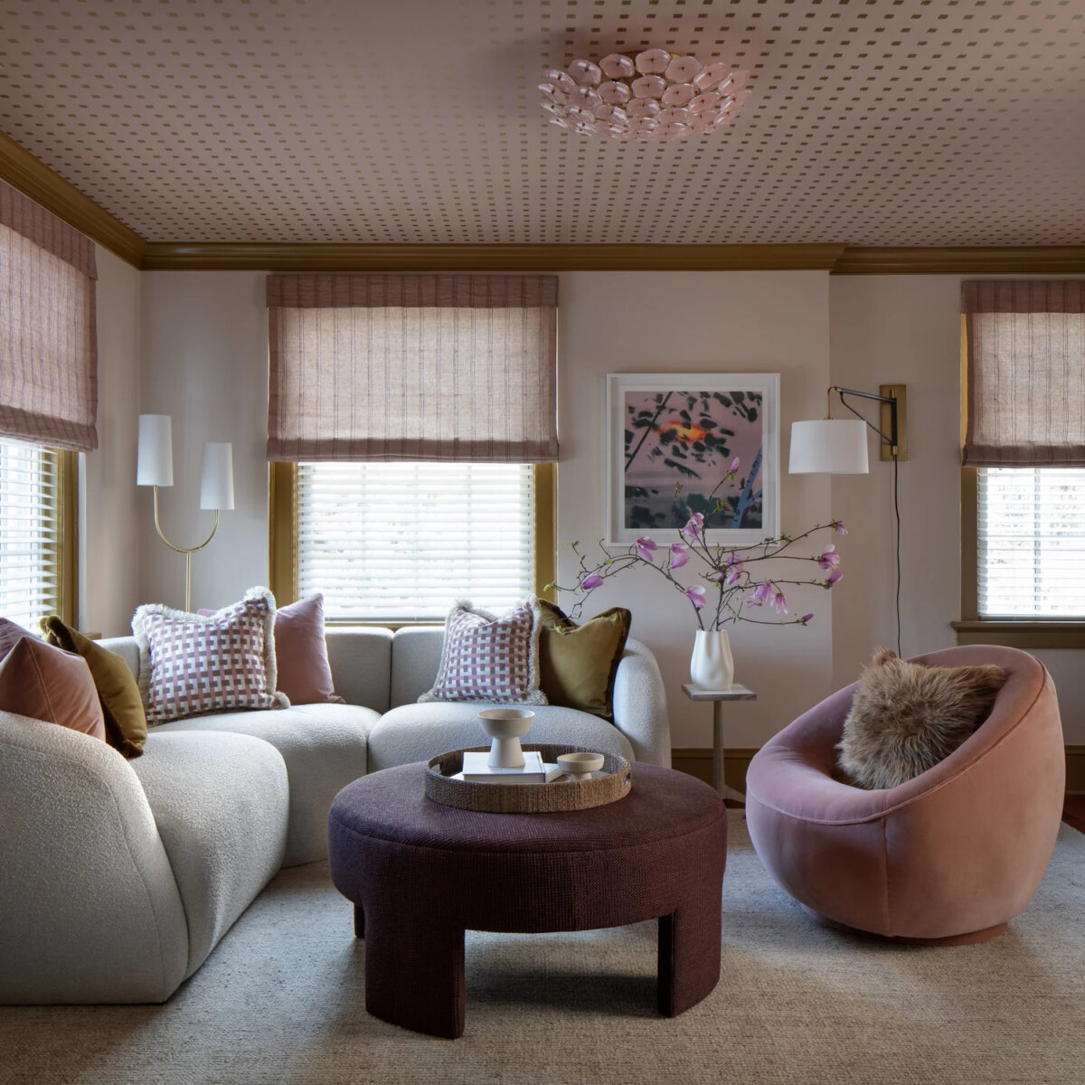 This Designer Wrote the Book on Creating Colorful Spaces — Literally! - 15 A modern designer living room with curved sofas, a round ottoman, and a blush accent chair. Neutral tones with pink and gold accents, and a vase with branches and wall art add decorative touches.