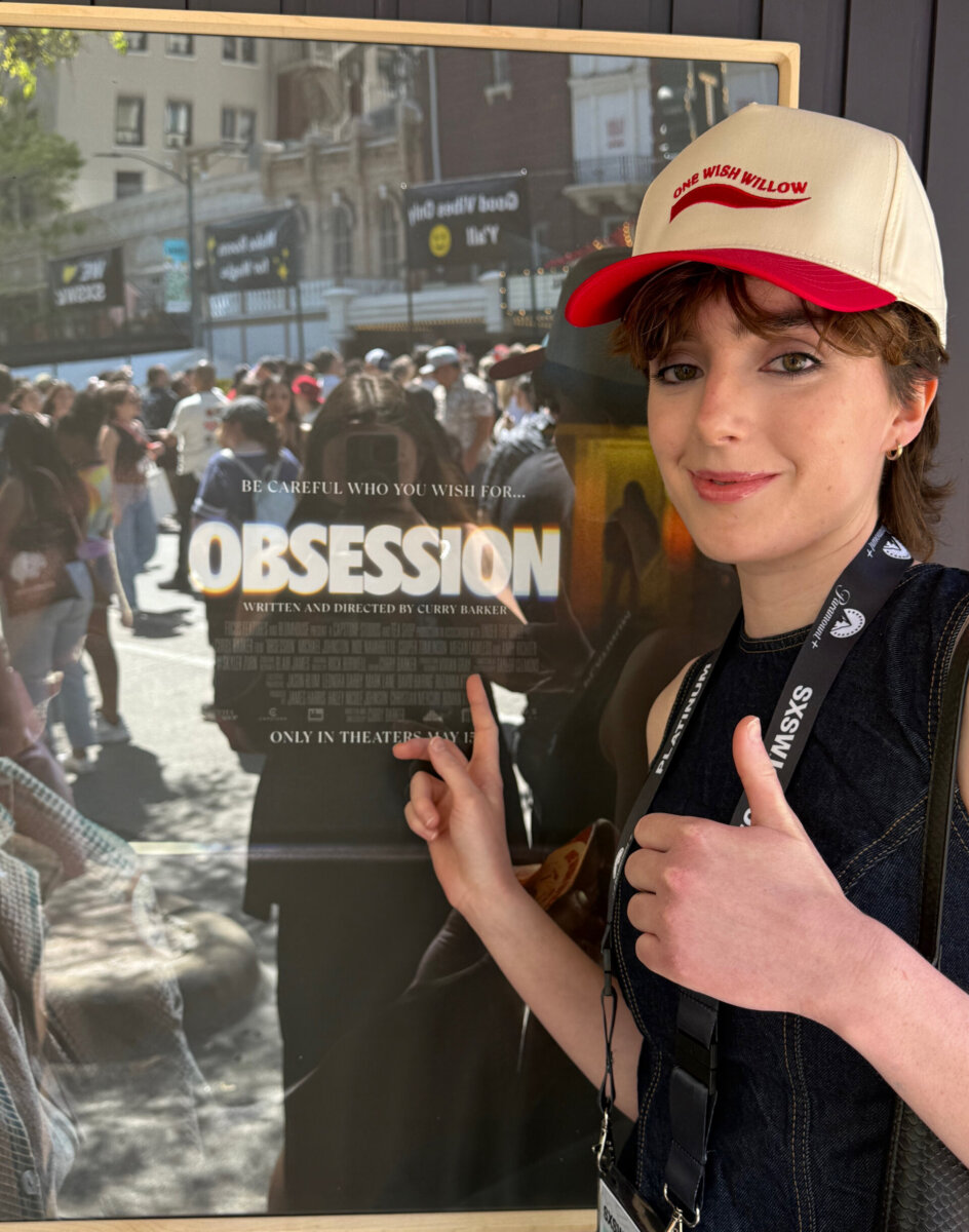 Meet the Memphis Designer Behind the Eerie World of "Obsession" - 5 A person wearing a hat and lanyard stands next to a movie poster for "Obsession" at the Memphian, pointing at the crafted title and giving a thumbs up.