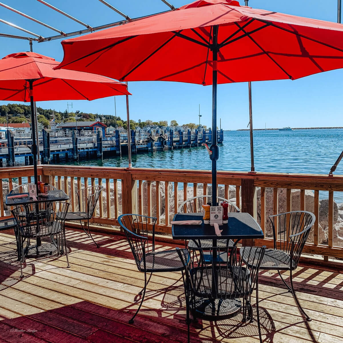 Midwest Weekend Getaway: My Mackinac Island Escape - 13 Outdoor dining area with black metal tables and chairs under red umbrellas, overlooking a marina with boats and clear blue water on a sunny day.