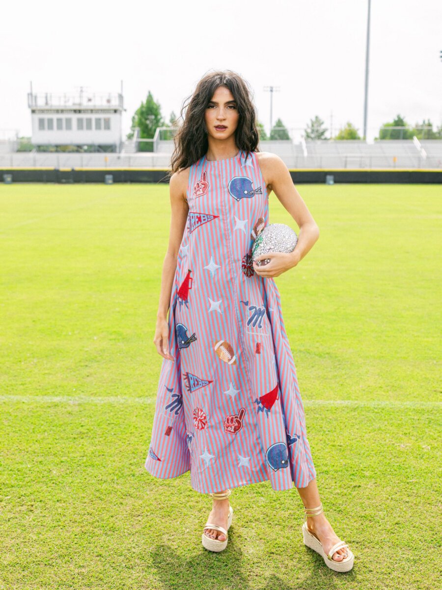 Southern Brands to Know for Football Season: Clothing, Decor & More - 3 Woman in a sleeveless, patterned dress stands on a grassy field, holding a sparkly clutch and wearing sandalsβperfectly showcasing her eye for accessories and effortless Southern brands style.