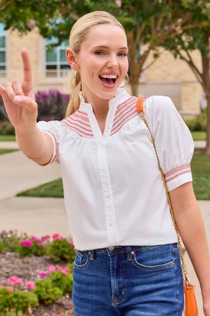 Southern Brands to Know for Football Season: Clothing, Decor & More - 4 Woman outdoors wearing a white blouse and blue jeans, smiling and holding up her hand with the index and pinky fingers extendedβshowcasing her excitement for football season accessories that complete her game-day look.