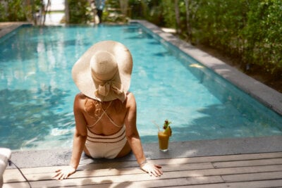 Woman in a sunhat and swimsuit sits at the edge of a swimming pool with her feet in the water and a tropical drink beside her, enjoying the sun safely outdoors instead of exposing herself to tanning bed risks.