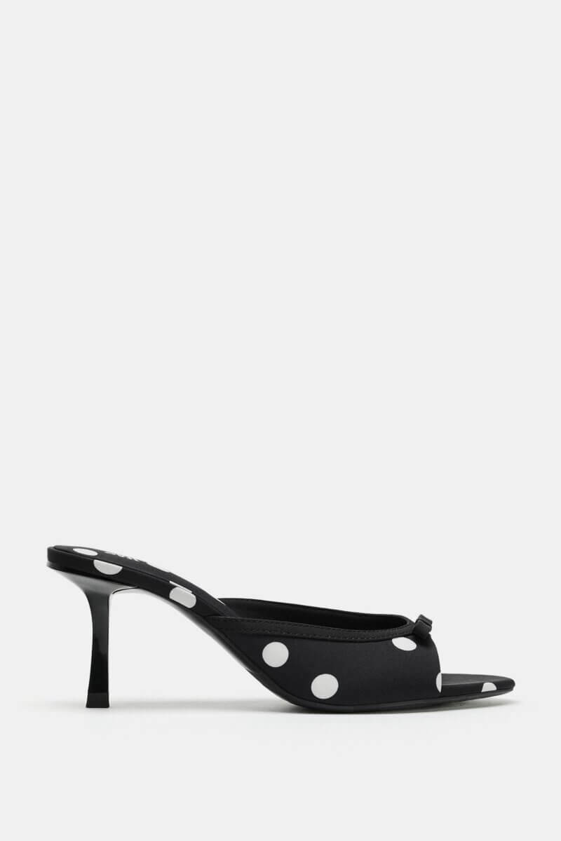 Black open-toe mule heel with white polka dots, a small bow detail on the front, and a mid-height stiletto heel—a playful pick that taps into timeless fashion trends. Shown on a plain white background.