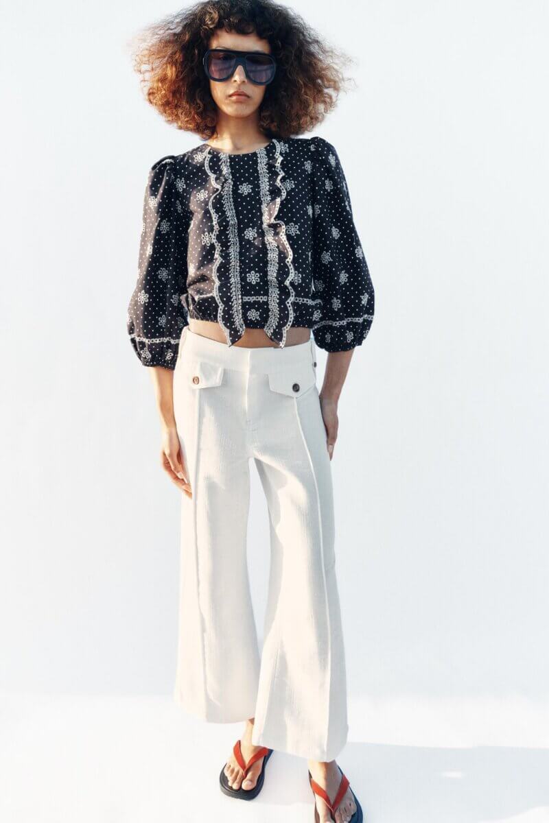 A person showcases polka dot fashion in a black patterned blouse, wide-leg white pants with buttons, red-and-black sandals, and sunglasses against a plain light background—perfect inspiration for how to wear polka dots.