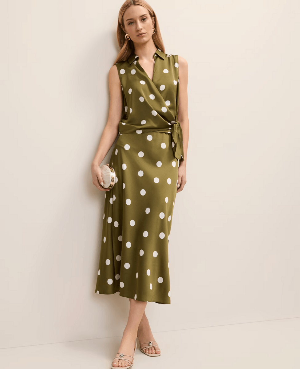 A woman stands against a wall wearing a sleeveless olive green wrap dress with white polka dots—showcasing how to wear one of this season's top fashion trends—holding a small clutch and wearing sandals.