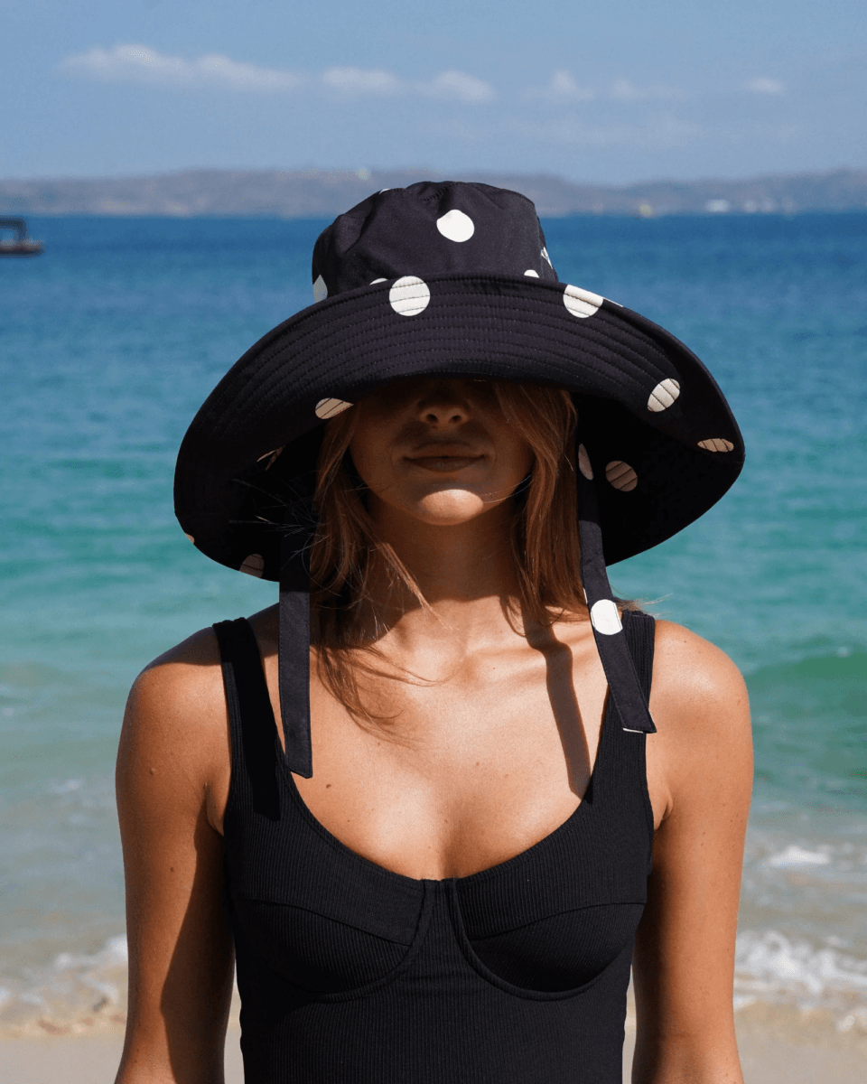 A woman wearing a large black polka dot sunhat and a black swimsuit stands on a beach, effortlessly showcasing how to wear one of this season’s top fashion trends against the backdrop of the ocean and distant shoreline.