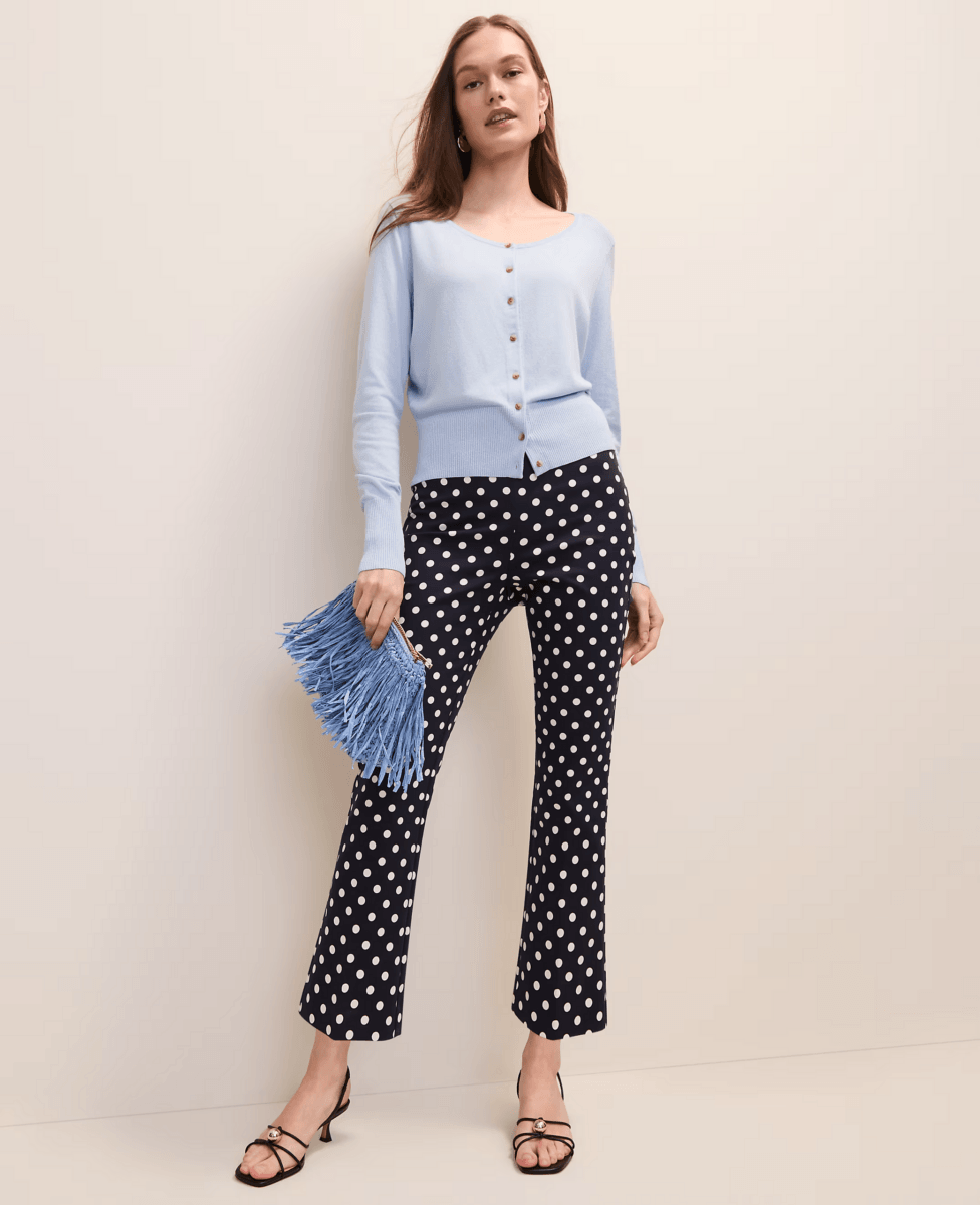 A woman showcases how to wear fashion trends with a light blue cardigan, navy polka dot pants, black heeled sandals, and a blue fringed clutch against a plain light background.
