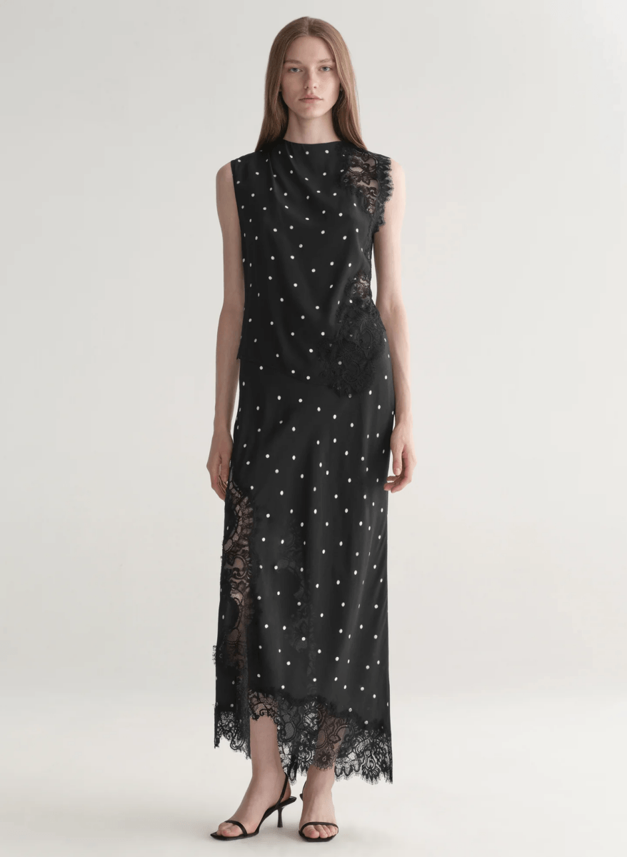 A woman stands facing forward wearing a sleeveless black dress with white polka dots, showcasing how to wear this timeless fashion trend, paired with lace detailing and black high-heeled sandals against a plain light background.