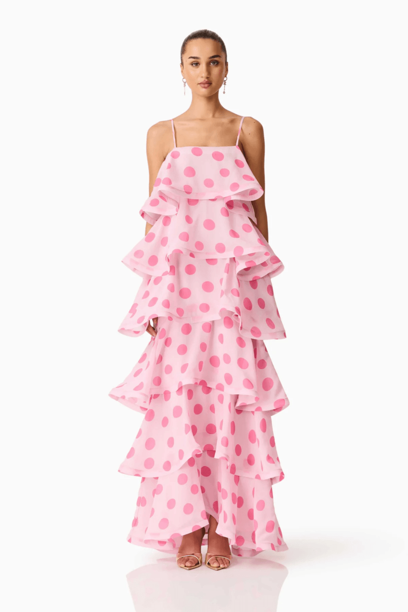 A woman stands wearing a sleeveless pink dress with large polka dots and multiple tiered ruffles, showcasing one of today’s top fashion trends, paired with open-toe heels against a plain white background.