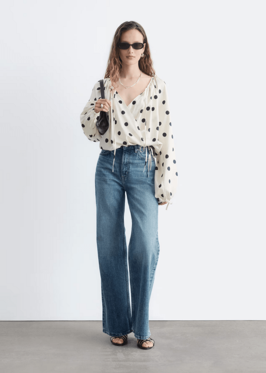 A person showcases how to wear Polka Dots, sporting a white polka dot blouse with blue high-waisted wide-leg jeans, black sandals, sunglasses, and a black handbag—capturing current fashion trends against a plain white background.