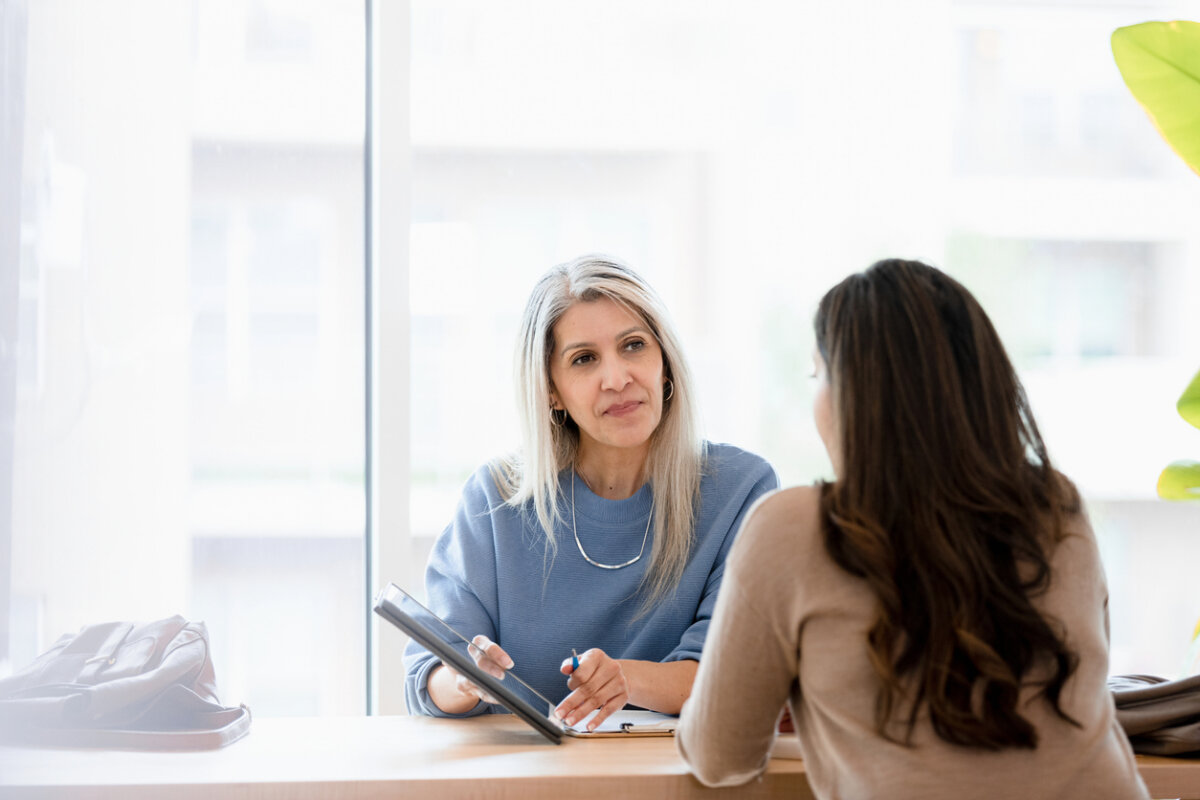 Why "Good" Finance Advice Doesn't Work for Everyone - 2 Two women sit across from each other at a table by a large window, one holding a tablet and pen, appearing to have a focused conversation as they discuss common financial misconceptions.