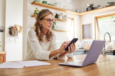Woman wearing glasses sits at a kitchen table, using a smartphone with one hand and a laptop in front of her, with papers and a coffee mug nearby.