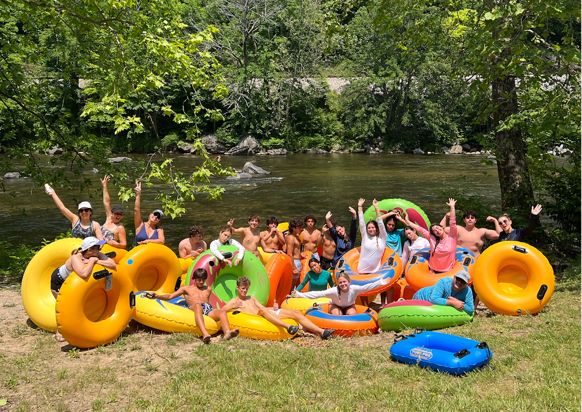 NC's Most Charming Mountain Road Trip Route: The Mayland Meander - 4 A group of people with colorful inflatable tubes pose and smile on grass by a river, surrounded by trees and greenery.