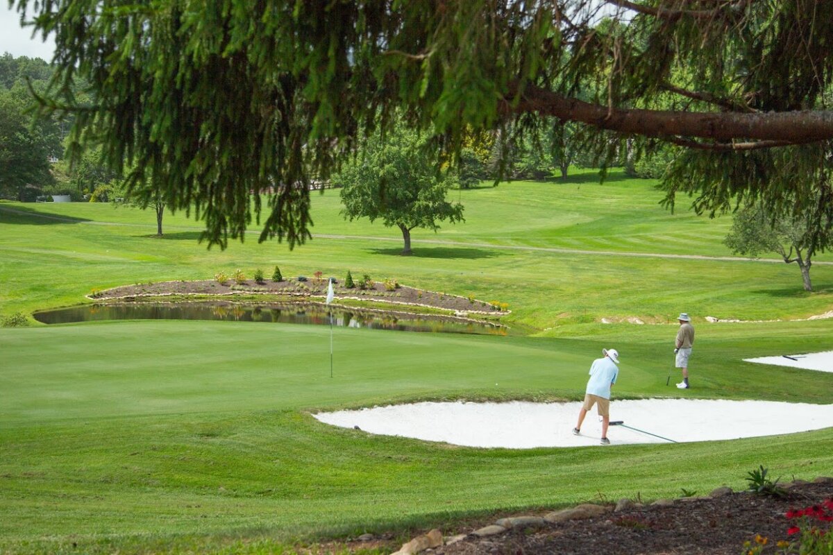 NC's Most Charming Mountain Road Trip Route: The Mayland Meander - 3 Two people are on a golf course along the scenic Mayland Meander in NC; one is raking a sand bunker near a green, while the other stands nearby. Trees and a pond are visible in the background, evoking the tranquil atmosphere of a mountain road trip.