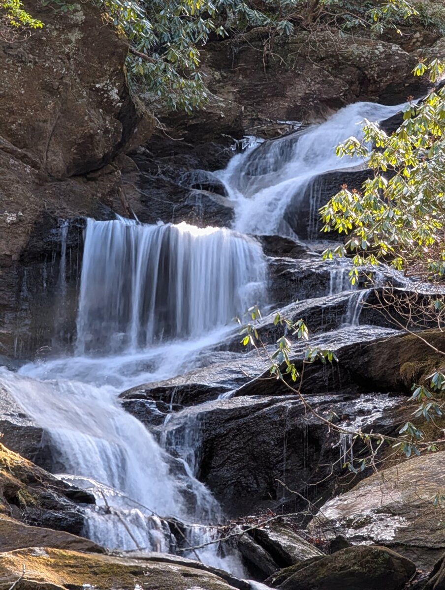 NC's Most Charming Mountain Road Trip Route: The Mayland Meander - 7 Multi-tiered waterfall cascades over dark rocks, surrounded by green foliage in a forested outdoor settingβa hidden gem often discovered along the charming mountain road of the Mayland Meander, perfect for an NC mountain road trip.