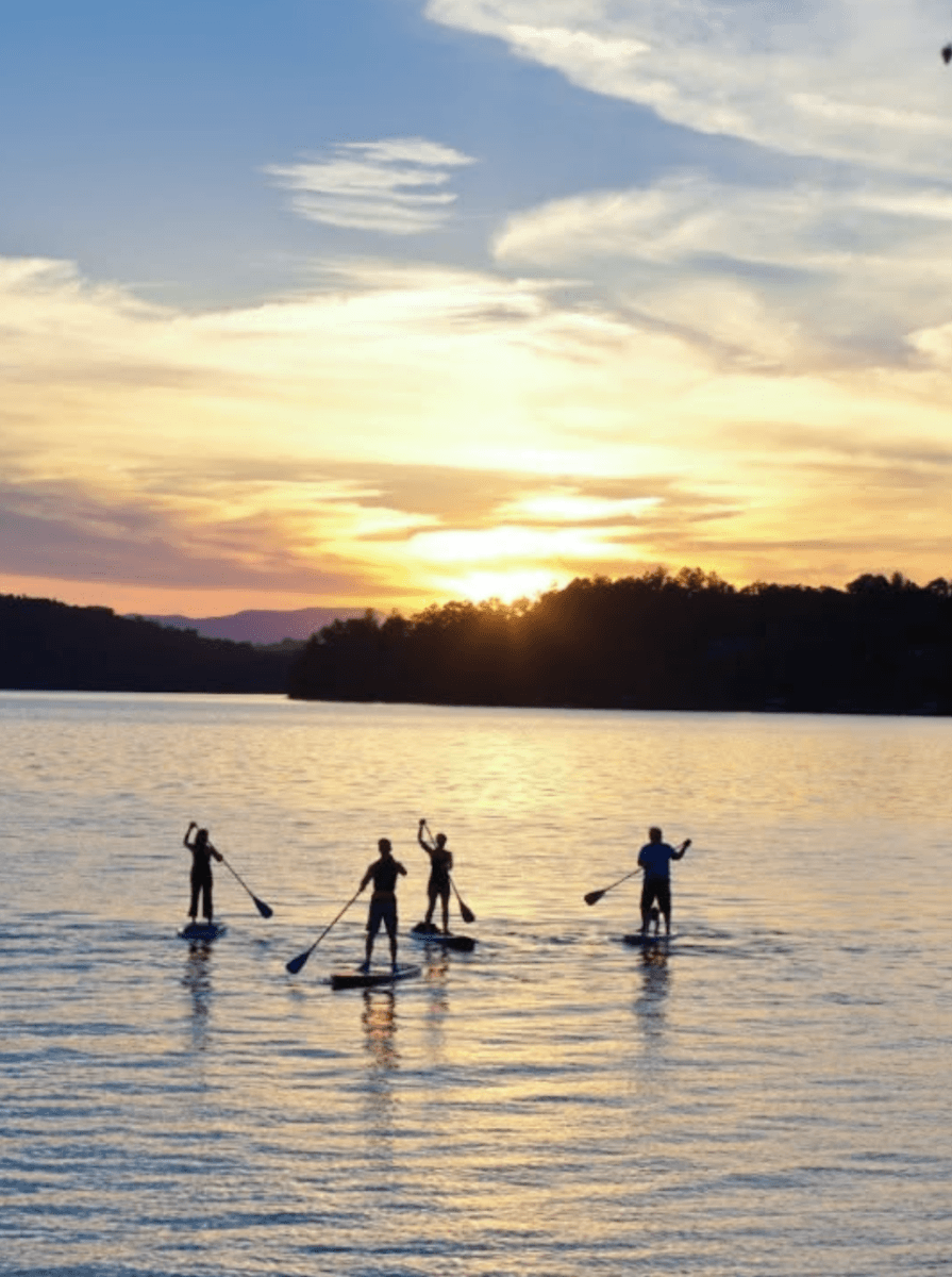 8 Easy Cool-Weather Escapes in the South & Beyond - 10 Four people stand-up paddleboarding on a calm lake at sunset, with silhouettes visible against the colorful sky and distant tree lineβan ideal scene for those seeking easy getaways or cool-weather escapes in the South.