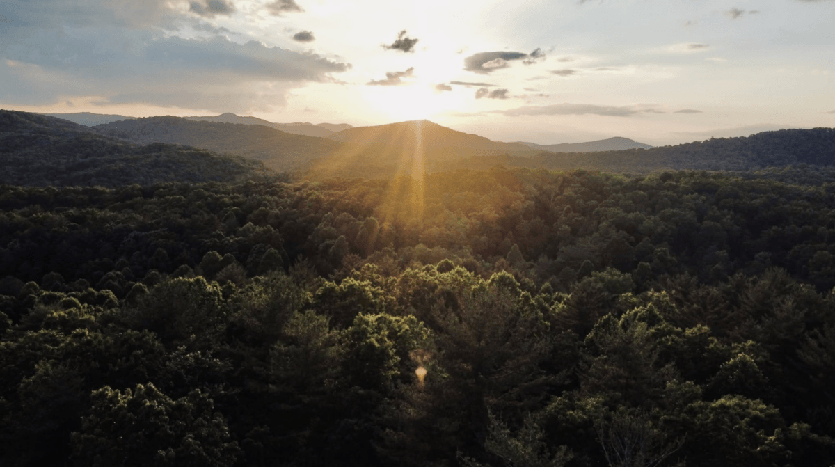 8 Easy Cool-Weather Escapes in the South & Beyond - 11 The sun sets over a forested mountain landscape in the South, casting rays of light through scattered clouds and illuminating the treetops belowβa perfect scene for those seeking cool-weather escapes and easy travel.