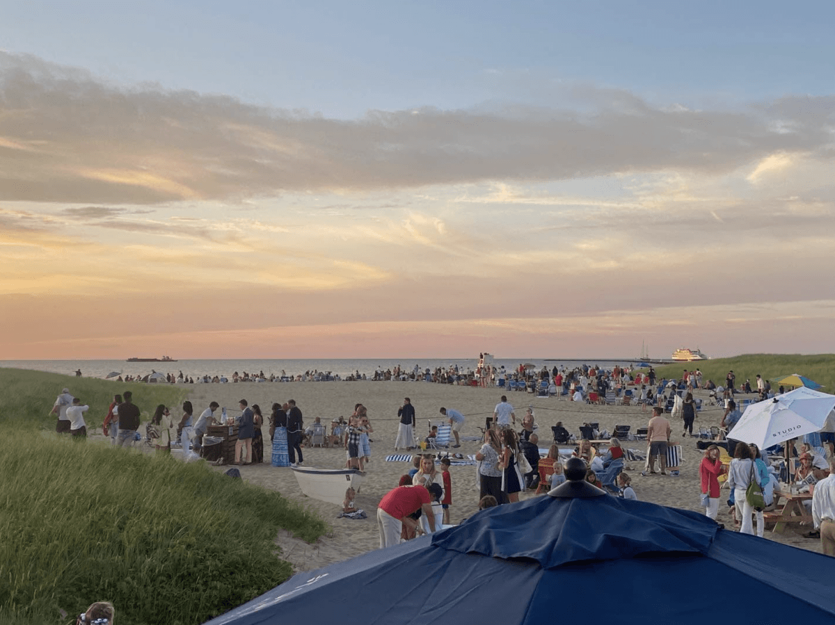 8 Easy Cool-Weather Escapes in the South & Beyond - 18 Crowd of people gathered on a sandy beach at sunset, with grassy dunes, umbrellas, and a ferry or ship visible in the distance.
