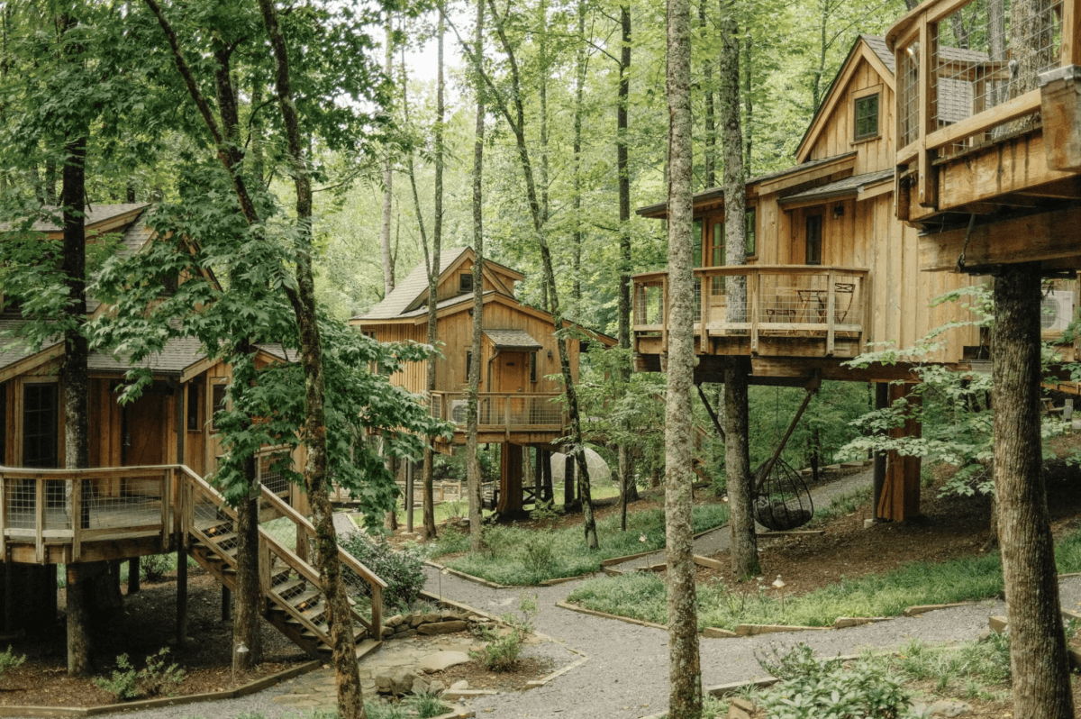 8 Easy Cool-Weather Escapes in the South & Beyond - 9 Cluster of wooden treehouses connected by walkways and stairs in a lush, green forest settingβan idyllic retreat for cool-weather escapes. Gravel paths wind between the buildings, inviting travelers seeking adventure and relaxation in the tranquil forests of the South.