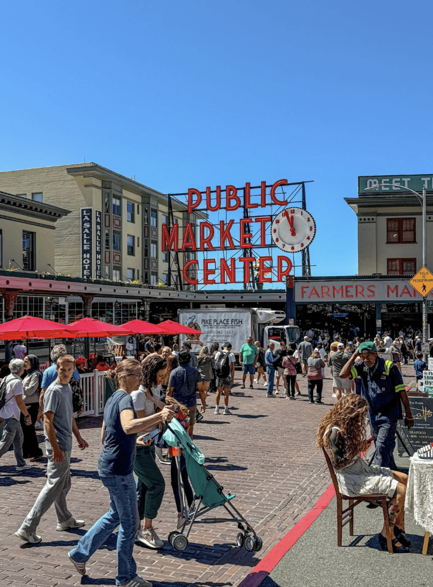 8 Easy Cool-Weather Escapes in the South & Beyond - 13 People walk near the entrance of Pike Place Market in Seattle on a sunny dayβone of the cityβs best cool-weather escapesβwith the red neon market sign and clock in the background.