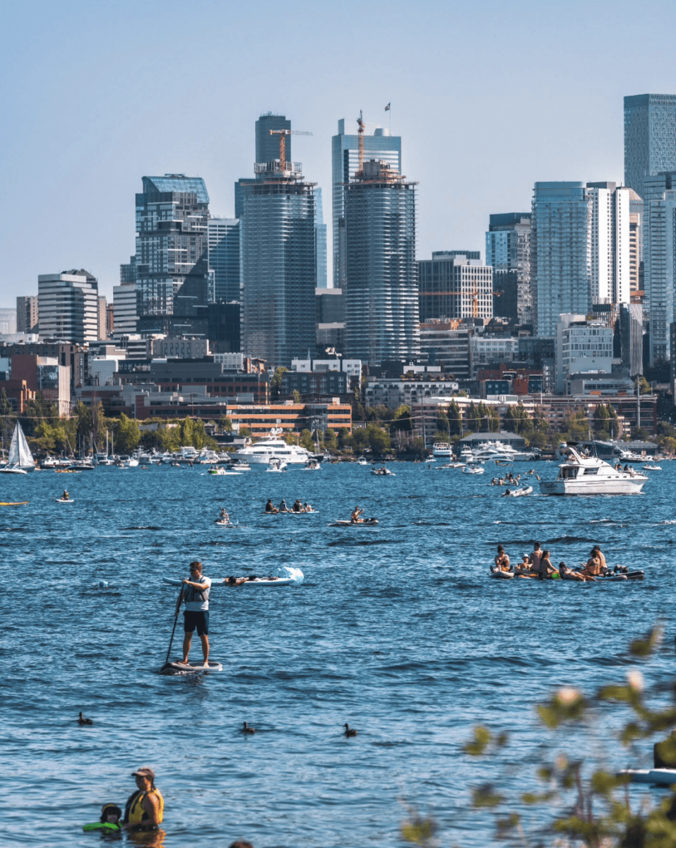 8 Easy Cool-Weather Escapes in the South & Beyond - 12 People paddle and relax on a lake with boats, paddle boards, and swimmers, with a city skyline and tall buildings in the background under a clear blue sky.