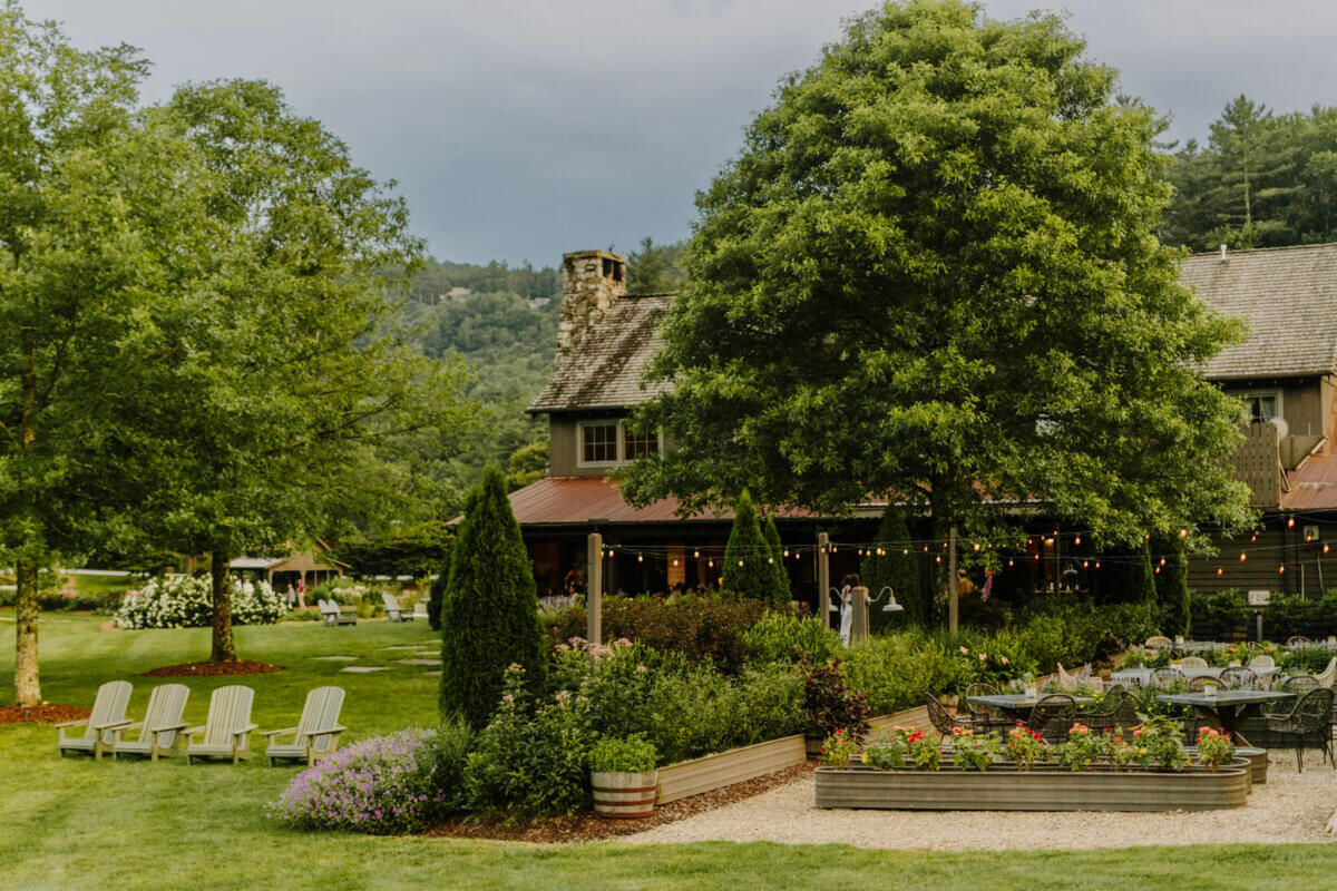 8 Easy Cool-Weather Escapes in the South & Beyond - 4 A rustic house with a stone chimney sits among lush gardens, string lights, outdoor seating, and Adirondack chairs, surrounded by greenery and treesβperfect for cool-weather escapes in the scenic South.