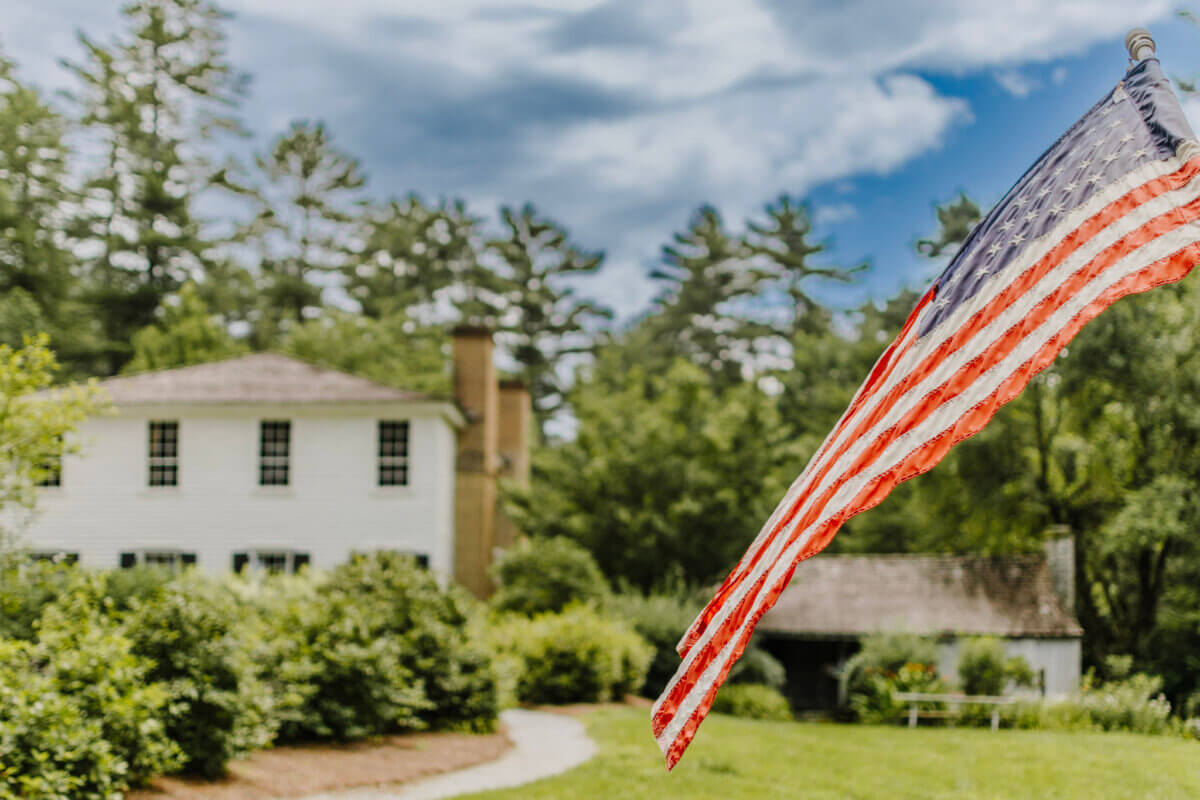 8 Easy Cool-Weather Escapes in the South & Beyond - 6 An American flag waves in the foreground with a white house, greenery, and trees under a partly cloudy sky in the backgroundβan inviting scene that captures the charm of cool-weather escapes ideal for travel in the South.