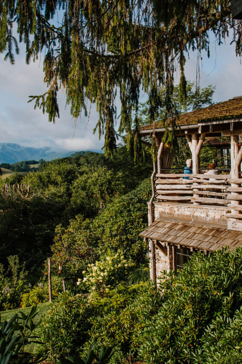 8 Easy Cool-Weather Escapes in the South & Beyond - 3 A rustic wooden cabin with a wraparound porch overlooks lush green trees and distant mountains under a partly cloudy sky, making it an ideal cool-weather escape in the South.