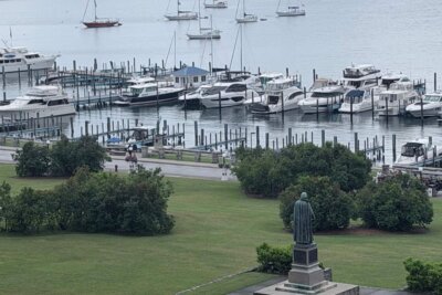 An elevated view of a marina filled with various motorboats and sailboats on Mackinac Island, situated behind a grassy park containing a bronze statue of a person viewed from behind.