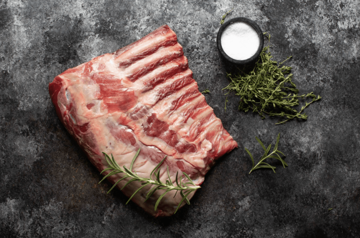Fire Up the Grill! 5 Best-Quality Meat Delivery Services in the South - 3 Raw rack of lamb with sprigs of rosemary and thyme lies on a dark surface next to a small bowl of salt, ready for the grill or your next meal from top meat delivery services in the South.