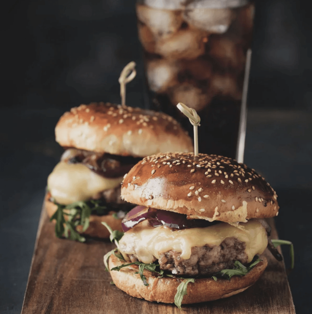 Fire Up the Grill! 5 Best-Quality Meat Delivery Services in the South - 6 Two cheeseburgers with lettuce and grilled onions sit on a wooden board, fresh off the grill, with a glass of iced cola in the backgroundβa classic taste of the South perfect for those who love hearty flavors or want to recreate this meal at home using trusted meat delivery services.